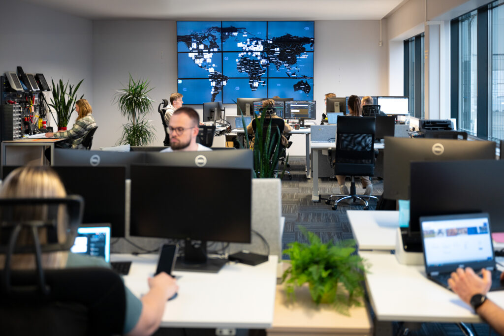 Office layout with people sitting behind tables, in background is a big wide screen with world map, reffering