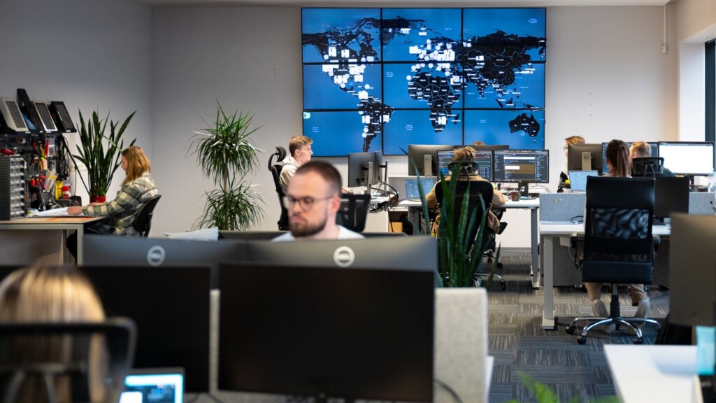 Wide view of the modern Cleveron Services operations center. Team members work at desks with multiple monitors, while a large video wall in the background displays a global map tracking active service locations. To the left, a technician manages a workspace equipped with hardware tools.